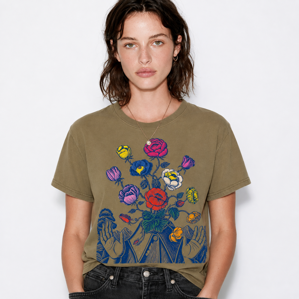 A woman wearing a muted olive green graphic t-shirt with a colorful illustration of flowers and hands, paired with black jeans, against a plain white background.