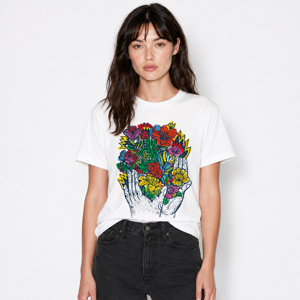 A woman modeling a white short-sleeve graphic t-shirt with a colorful illustration of hands holding a vibrant bouquet of flowers, paired with black jeans, against a plain white background.