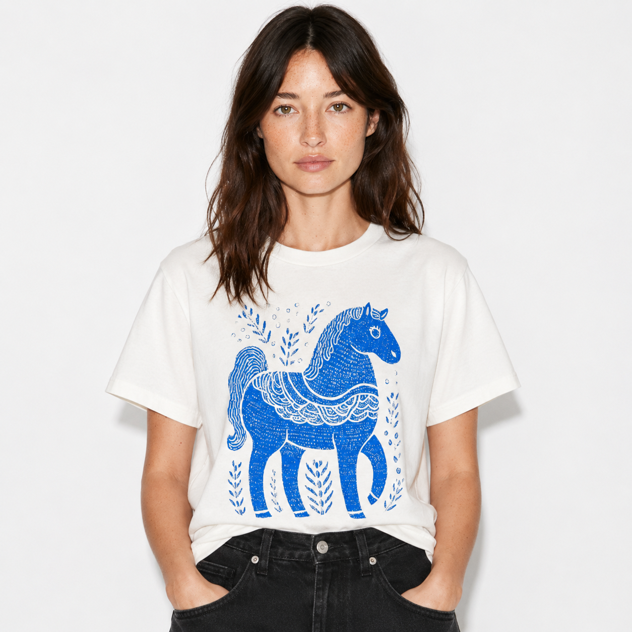 A woman wearing a white short-sleeve t-shirt with a blue folk-art horse graphic, paired with black jeans, against a plain white background.