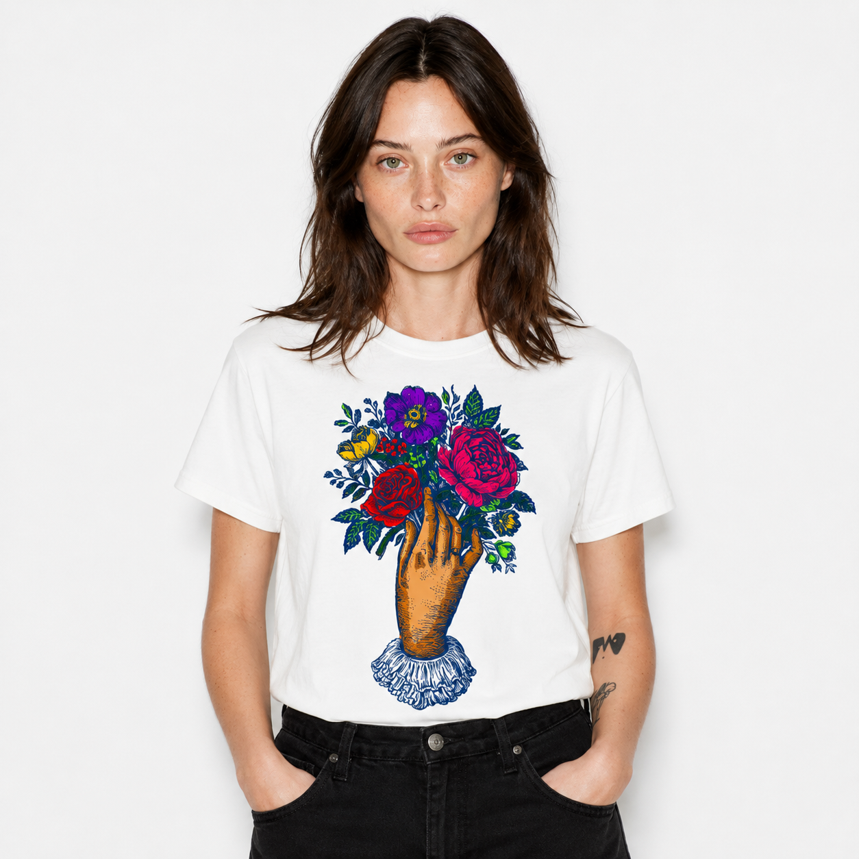 A woman wearing a white short-sleeve t-shirt with a colorful graphic of a hand holding a bouquet of red, pink, and purple flowers, paired with black jeans, against a plain white background.