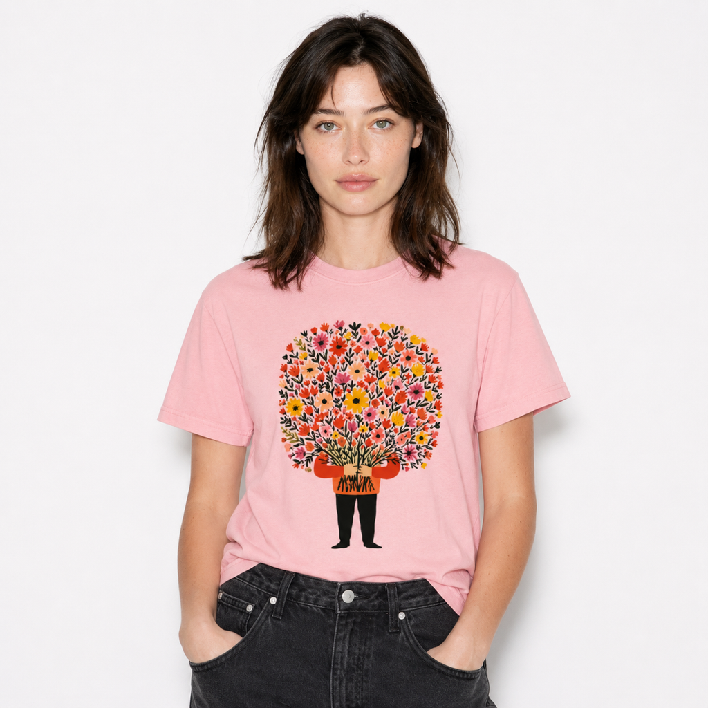 A woman wearing a light pink crewneck t-shirt with a colorful graphic of a person holding a vibrant bouquet of flowers. She is paired with dark gray jeans, standing against a clean white background.