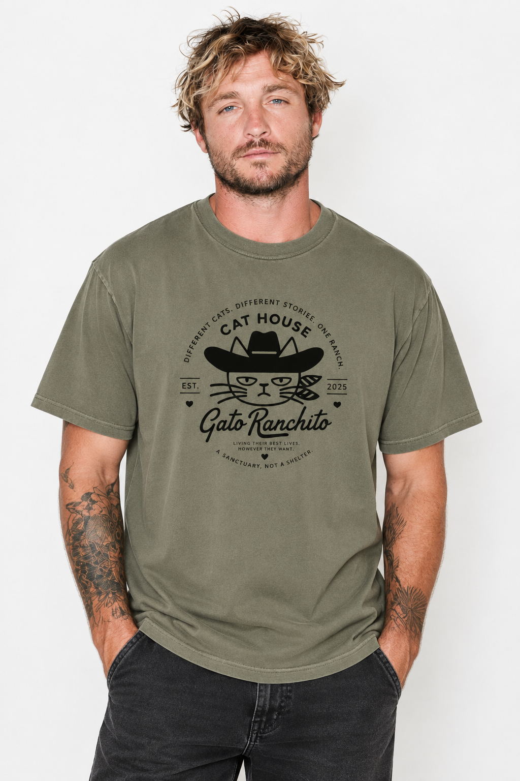A man wearing an olive green graphic t-shirt with a black cat rancher design, paired with dark jeans, against a plain light gray background.