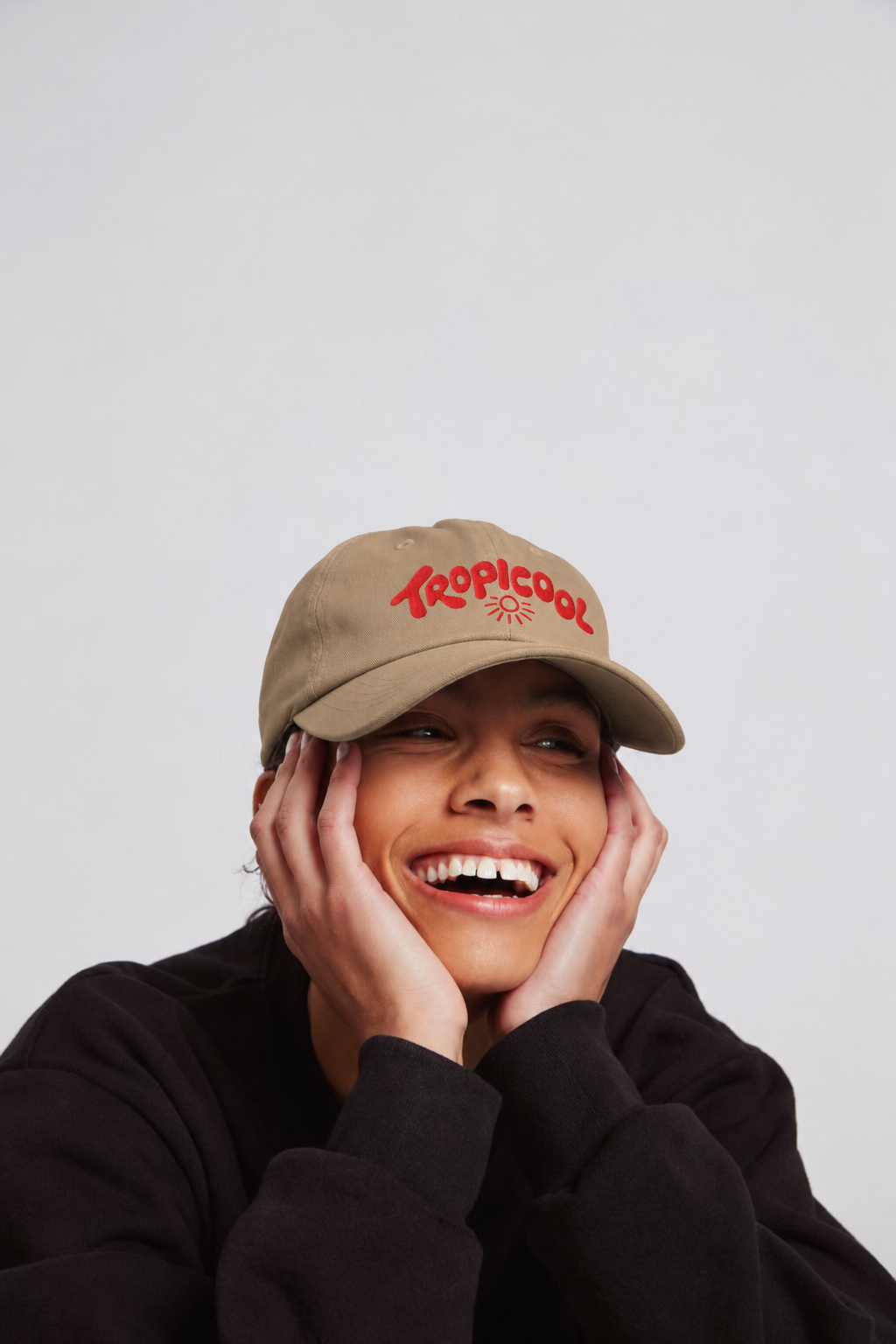 A smiling woman wearing a tan baseball cap with red embroidered 'Tropicool' branding, posing with her hands on her cheeks against a light gray background.