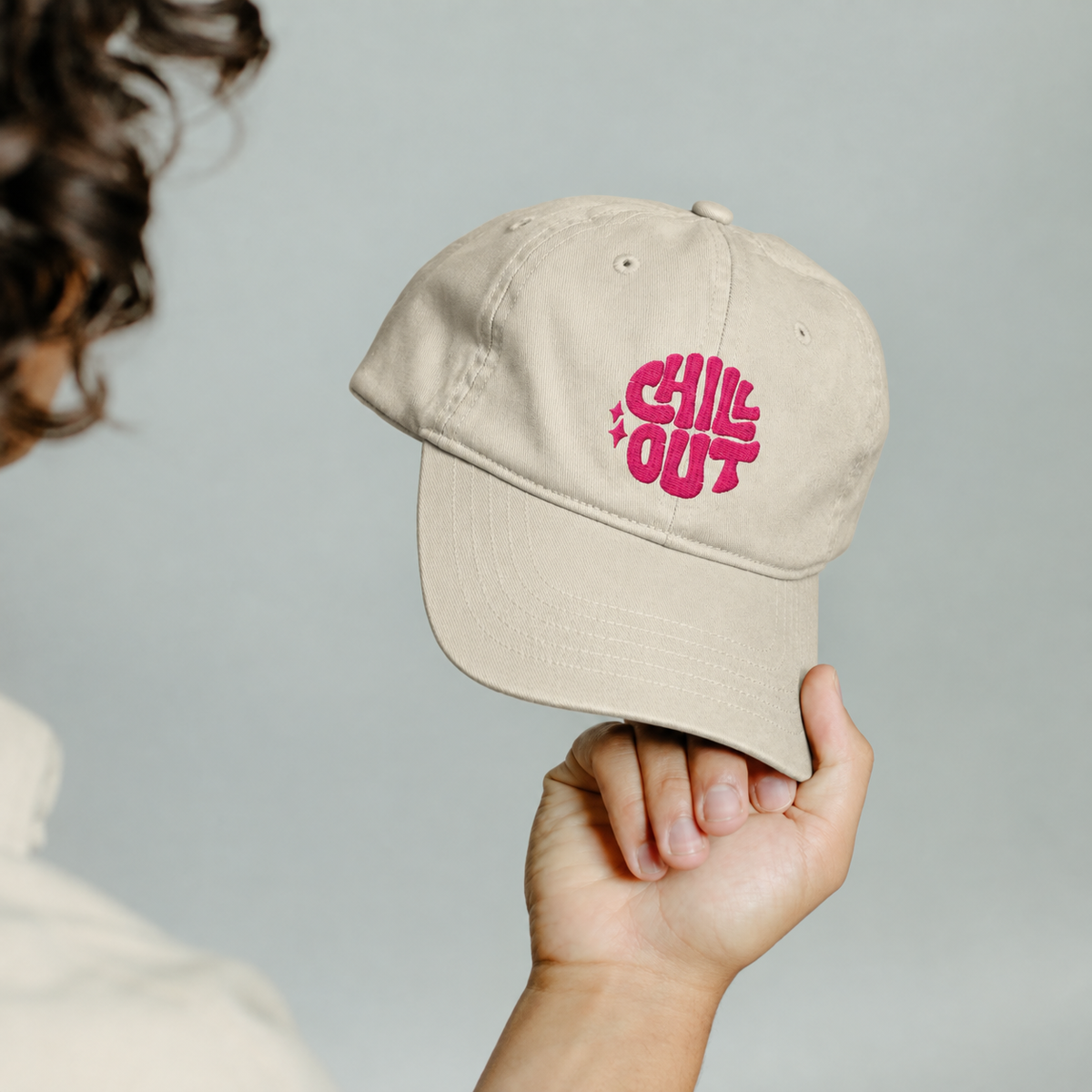 A person holds a beige baseball cap with a pink embroidered 'CHILL OUT' graphic on the front, shown against a soft light gray background.