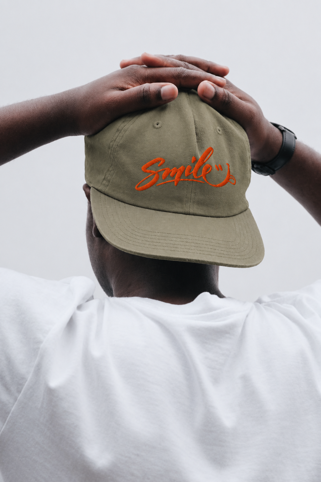 A person viewed from behind wearing an olive green baseball cap with orange embroidered text reading 'Smile' and a white T-shirt, against a plain light gray background.