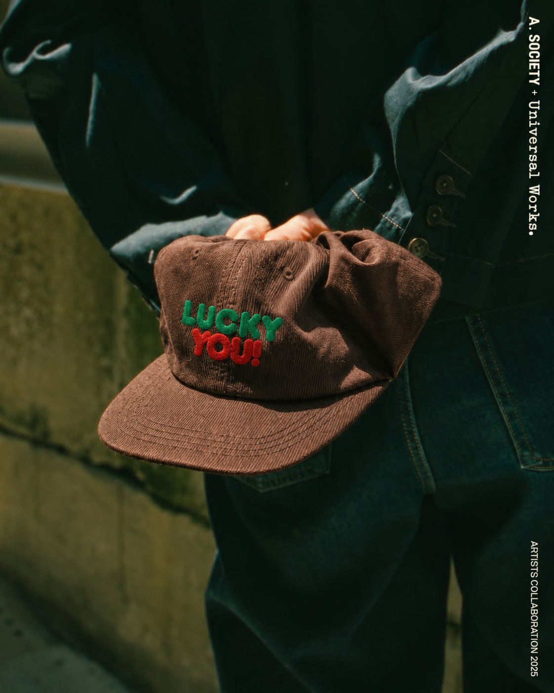 A person wearing a dark denim jacket and jeans holds a brown baseball cap with 'LUCKY YOU!' embroidered on the front in green and red. The photo is shot in warm, natural light against a concrete wall, with vertical text on the right side.