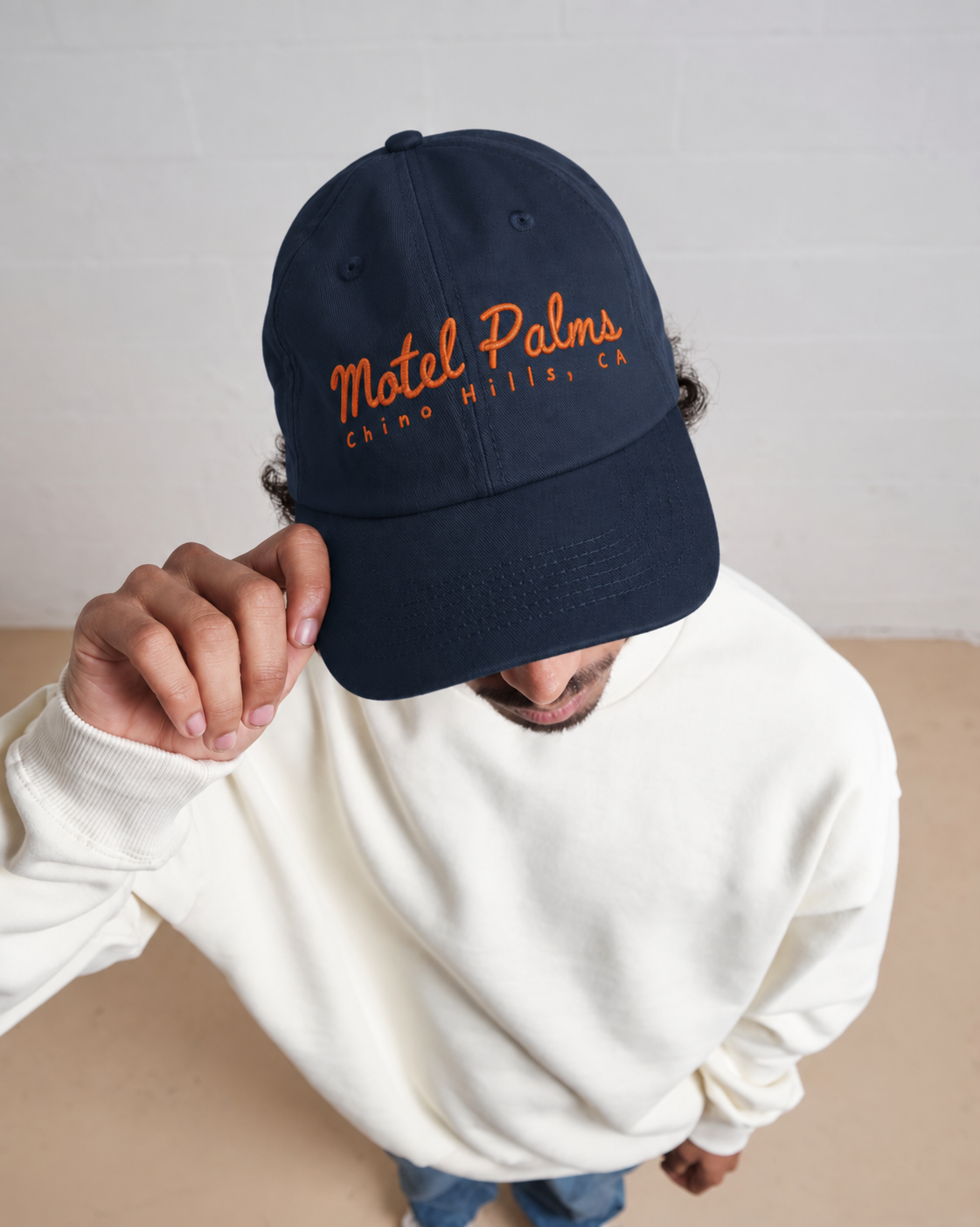 A man wearing a navy blue baseball cap with orange embroidered text reading 'Motel Palms, Carona Hills, CA', paired with a cream sweatshirt and blue jeans, photographed against a light gray brick wall.