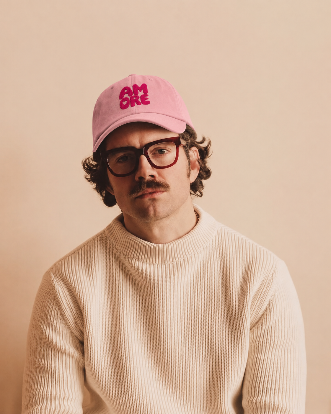 A man wearing a pink baseball cap with 'SMORE' in red text, red-framed glasses, and a cream ribbed sweater, photographed against a warm beige background.