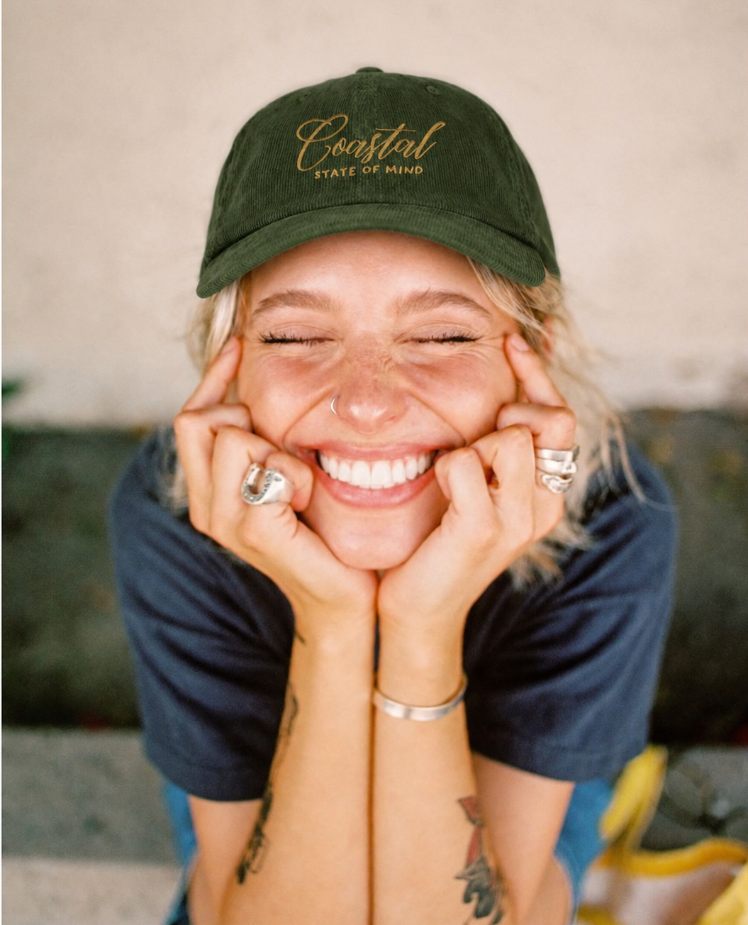 A smiling woman with closed eyes rests her head on her hands, wearing a dark green baseball cap with gold embroidered text reading 'Coastal State of Mind' and a navy blue t-shirt.