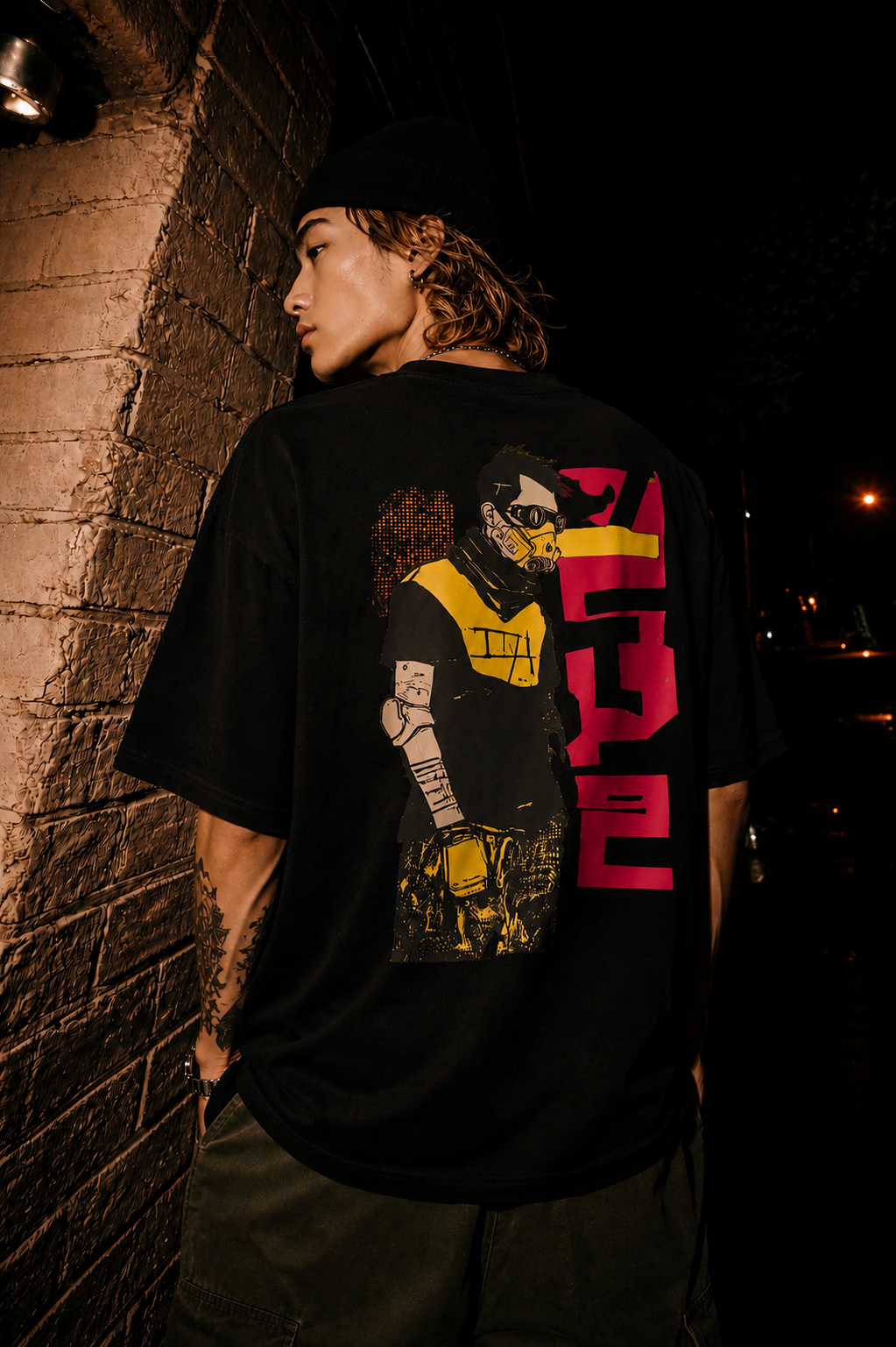 A person wearing a black oversized graphic T-shirt stands against a dimly lit brick wall at night, showing the back of the shirt with a large illustrated print in yellow, pink, and black.