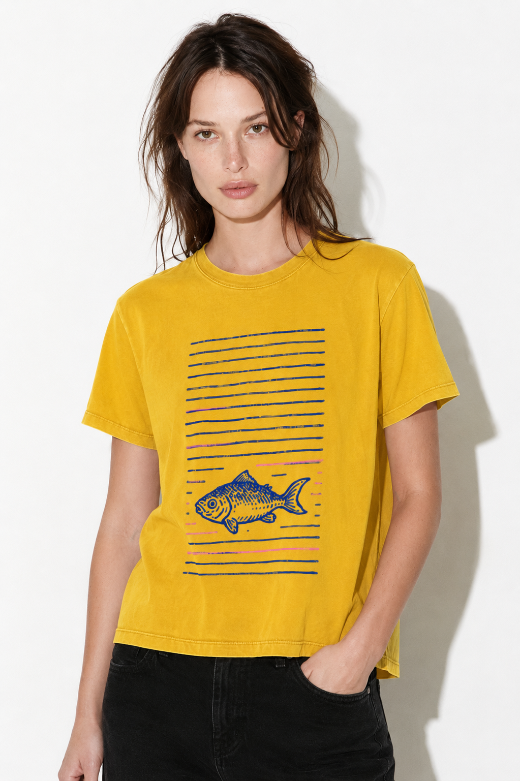 A woman modeling a yellow short-sleeve graphic t-shirt with a blue striped pattern and a fish illustration on the front, paired with black jeans against a white studio background.