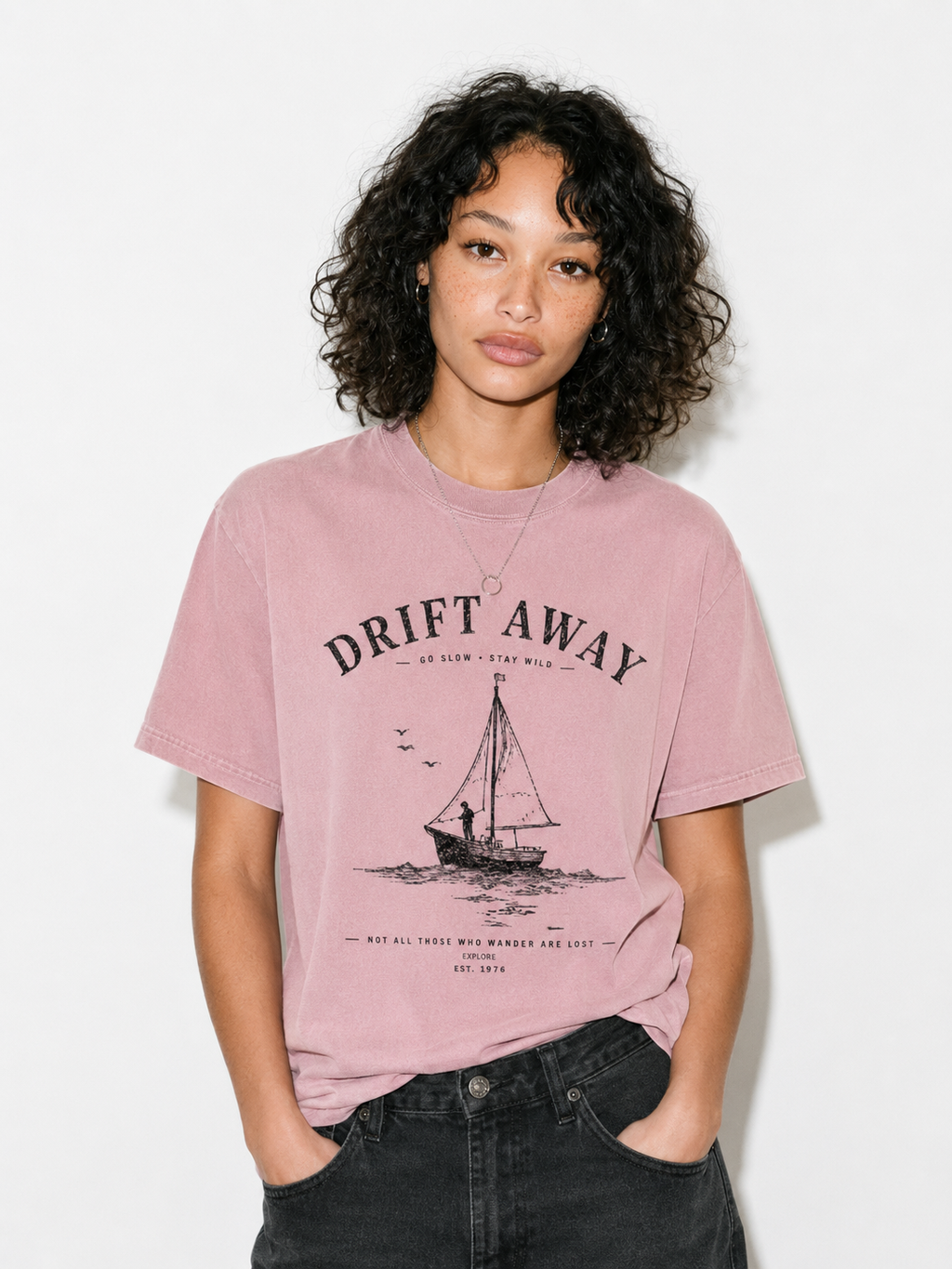 A woman with curly dark hair wearing a dusty pink graphic t-shirt with a black sailboat print and the text 'DRIFT AWAY'. She is paired with dark gray jeans, standing against a clean white background.