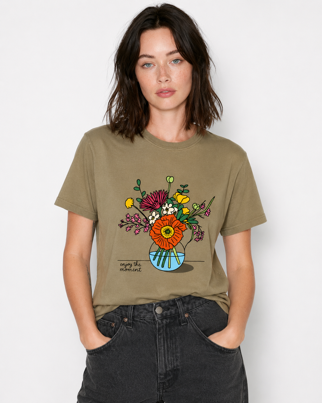 A woman wearing an olive green graphic t-shirt with a colorful flower illustration on the front, paired with dark gray jeans, against a plain light gray background.