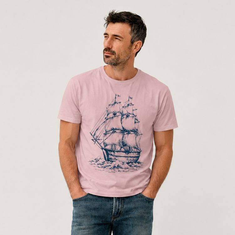 A man wearing a light pink crewneck t-shirt with a dark blue sailing ship graphic, paired with blue jeans and white sneakers, standing against a plain light gray background.