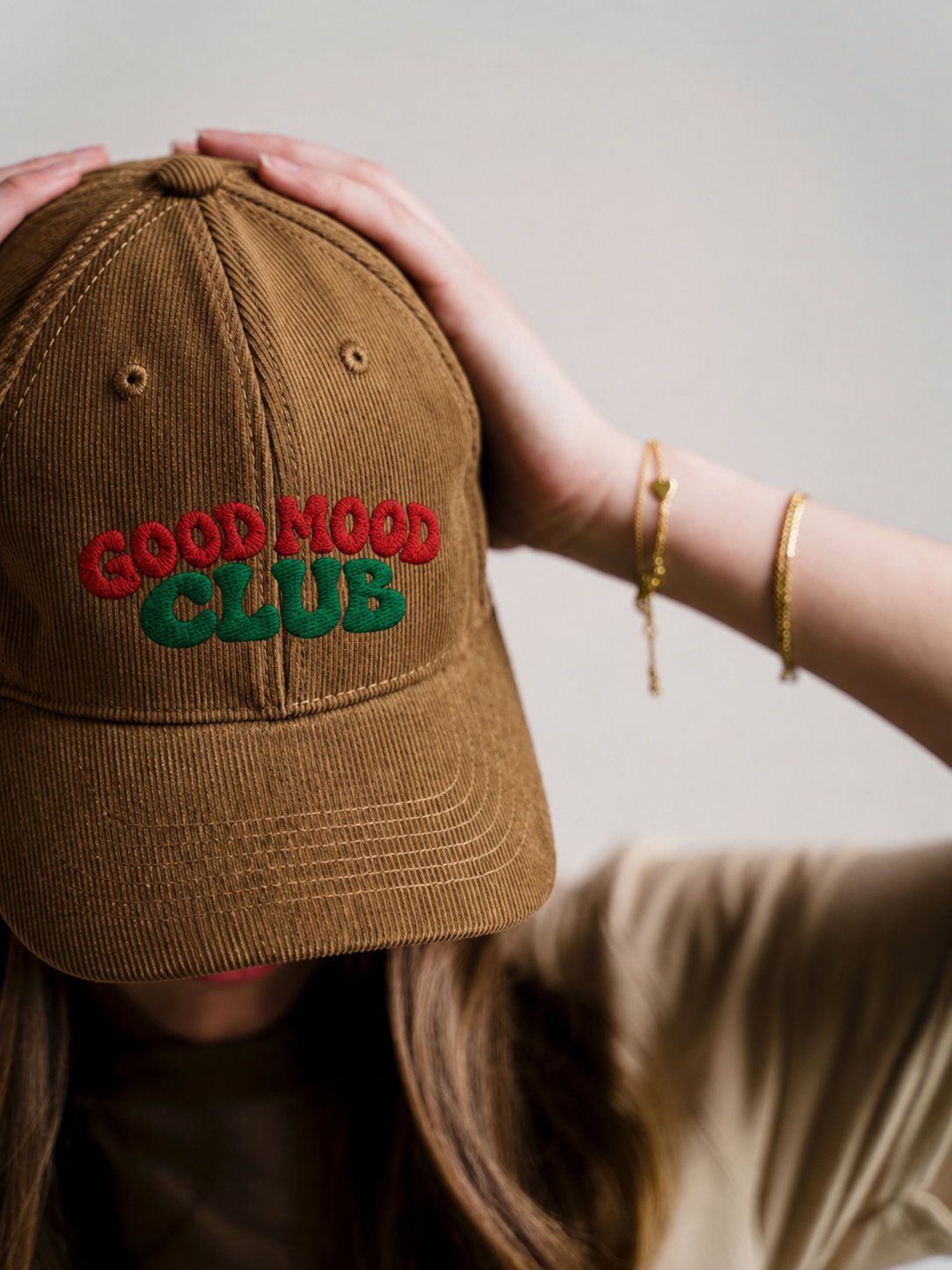 A person wearing a brown corduroy baseball cap with 'GOOD MOOD CLUB' embroidered on the front in red and green, with their hands holding the top of the cap against a neutral light gray background.