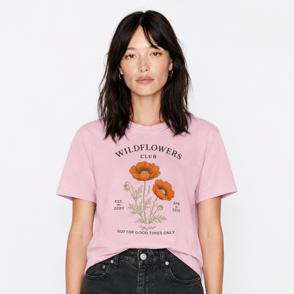 A woman wearing a light pink graphic t-shirt with orange poppy illustrations and the text 'WILDFLOWERS CLUB', paired with black jeans, against a plain white background.