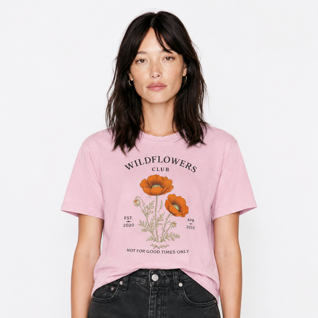 A woman wearing a light pink graphic t-shirt with orange poppy illustrations and the text 'WILDFLOWERS CLUB', paired with black jeans, against a plain white background.