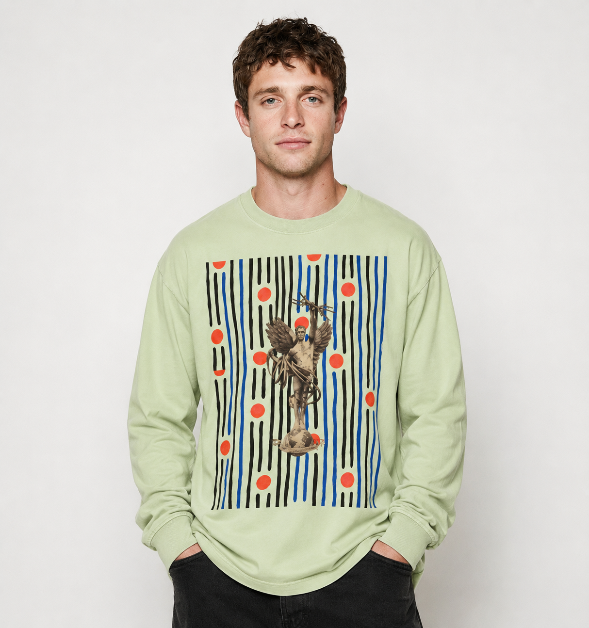 A man wearing a light sage green long-sleeve t-shirt with a large graphic print on the front. The graphic features vertical black and blue stripes, orange polka dots, and a central illustration of a bird with a blue hat.