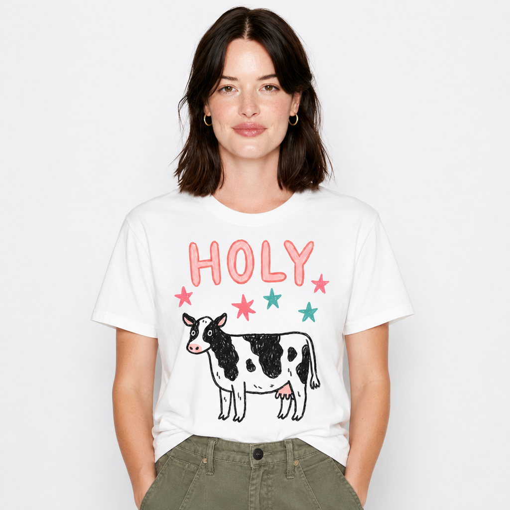 A woman wearing a white graphic t-shirt with a black and white cow and the word 'HOLY' in pink and teal, paired with olive green pants, against a light gray background.