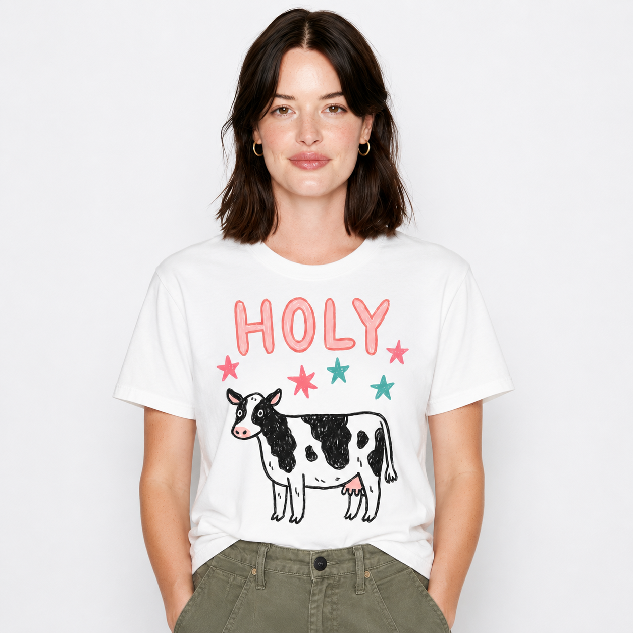 A woman wearing a white graphic t-shirt with a black and white cow and the word 'HOLY' in pink and teal, paired with olive green pants, against a light gray background.