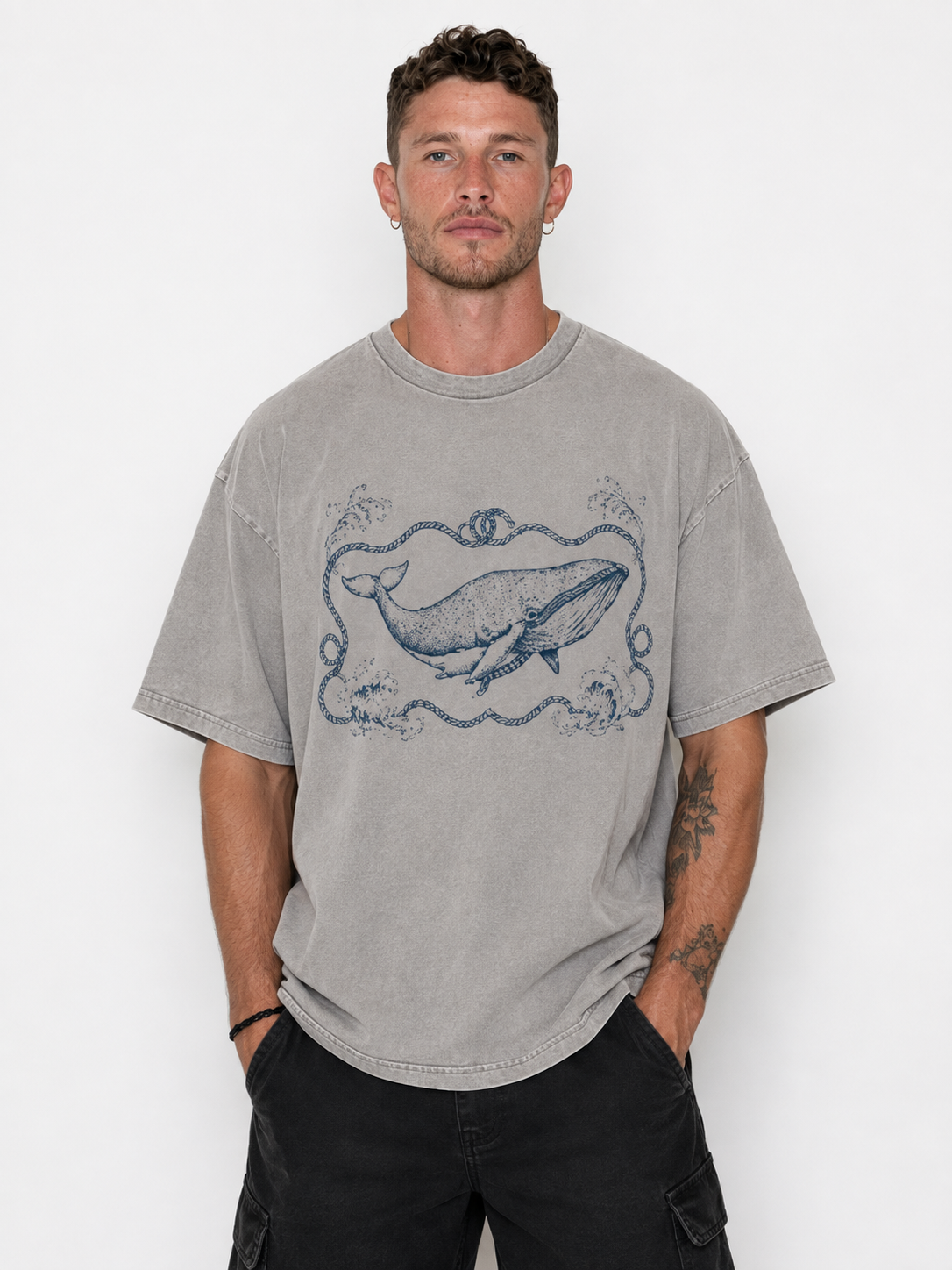 A man wearing a gray washed t-shirt with a blue whale graphic on the front, paired with black cargo pants, standing against a white studio background.