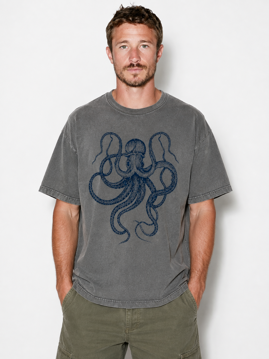 A man wearing a gray washed t-shirt with a dark blue octopus graphic on the front, paired with olive green cargo pants, standing against a light gray background.