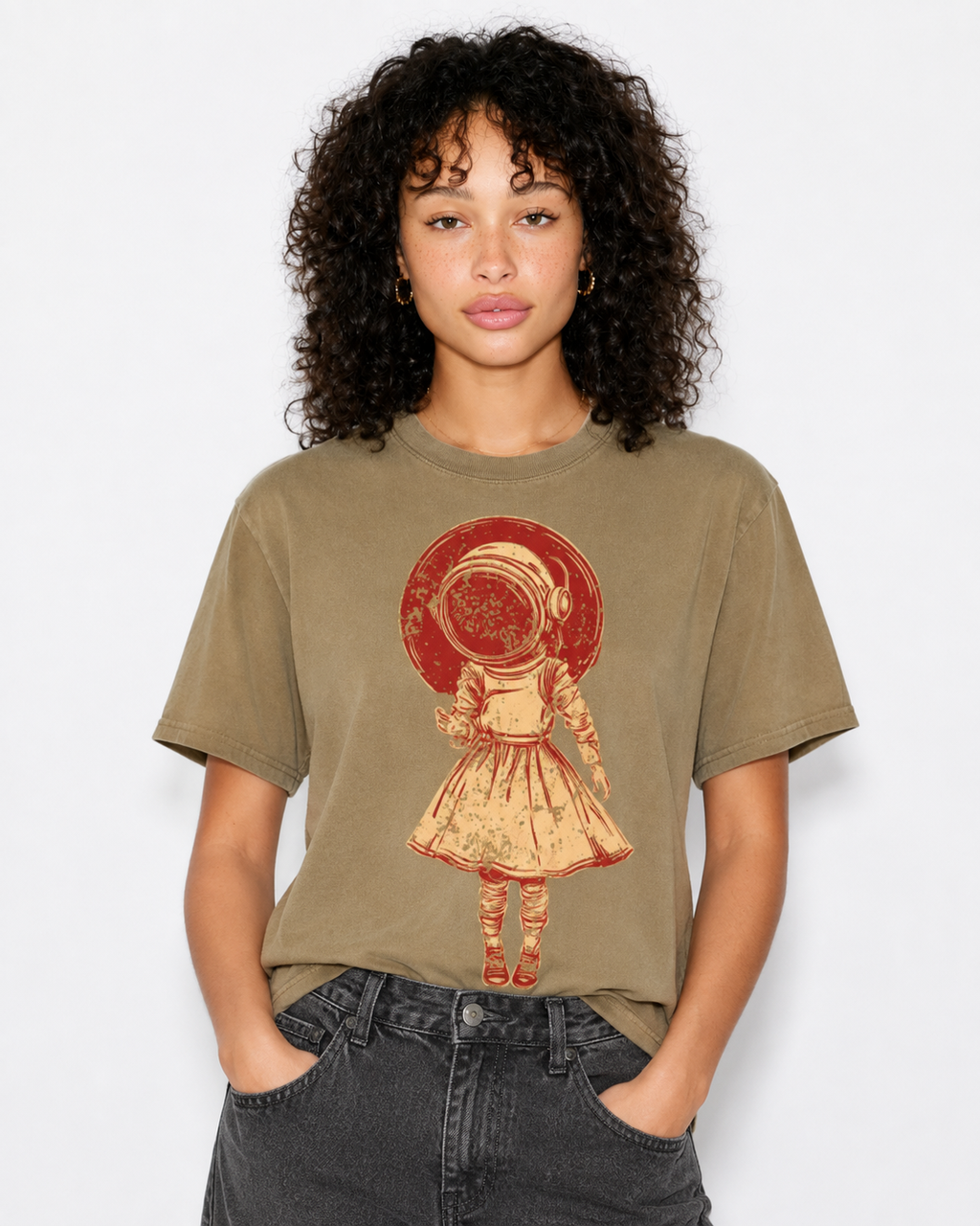A woman with curly dark hair wearing a faded olive-brown graphic t-shirt featuring a retro illustration of an astronaut girl in a red helmet, paired with dark gray jeans, against a plain light gray background.