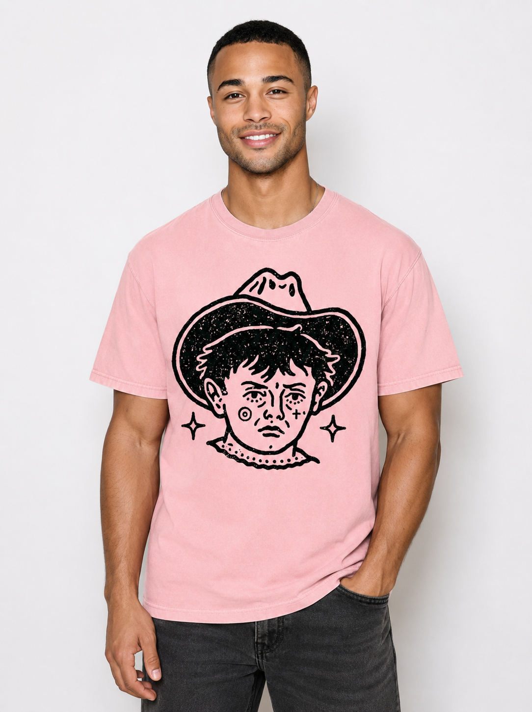 A man wearing a light pink crewneck t-shirt with a black graphic print of a face wearing a cowboy hat, paired with dark gray jeans, against a plain light gray background.