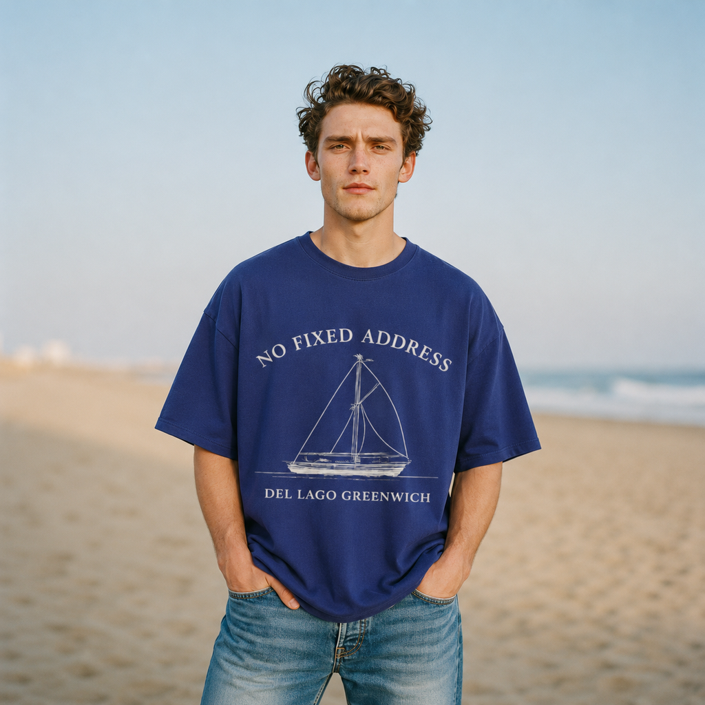 A man standing on a sandy beach wearing an oversized navy blue t-shirt with a white sailboat graphic and the text 'NO FIXED ADDRESS DEL LAGO GREENWICH'.