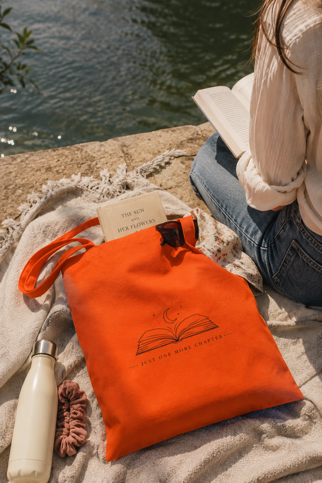 An orange canvas tote bag with a black line art graphic of an open book and the words 'ONE MORE CHAPTER' sits on a beige blanket next to a person sitting by the water, with a white bottle and a pink scrunchie nearby.