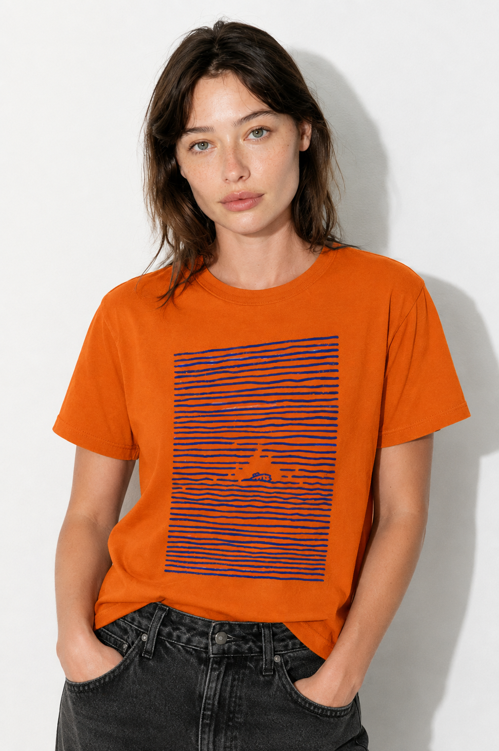 A woman wearing a bright orange crewneck t-shirt with a graphic of horizontal navy and red stripes on the front, paired with dark gray jeans, standing against a white wall.
