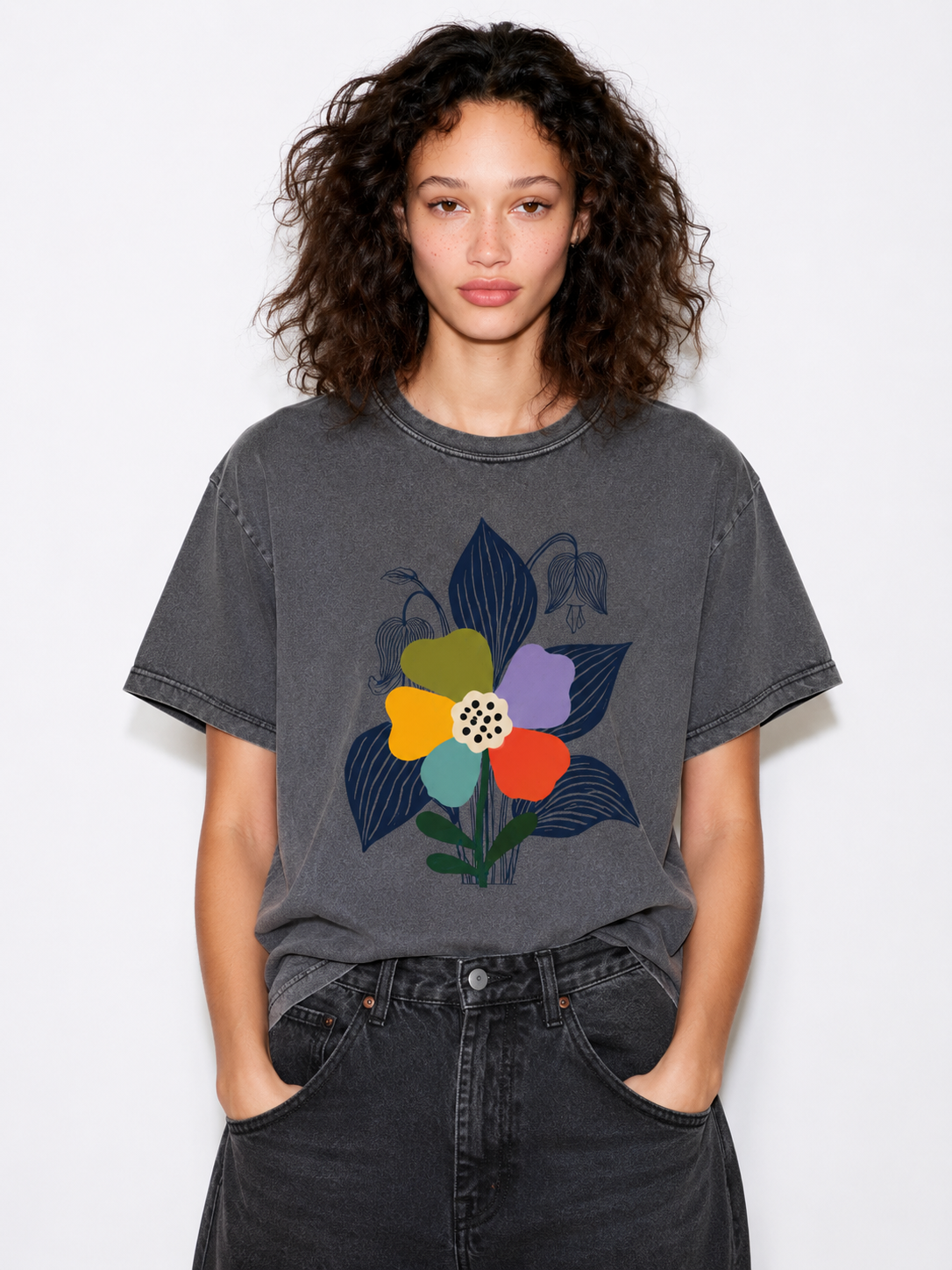 A woman with curly dark hair wearing a faded charcoal gray t-shirt with a colorful graphic of stylized flowers, paired with dark gray jeans, against a plain white background.