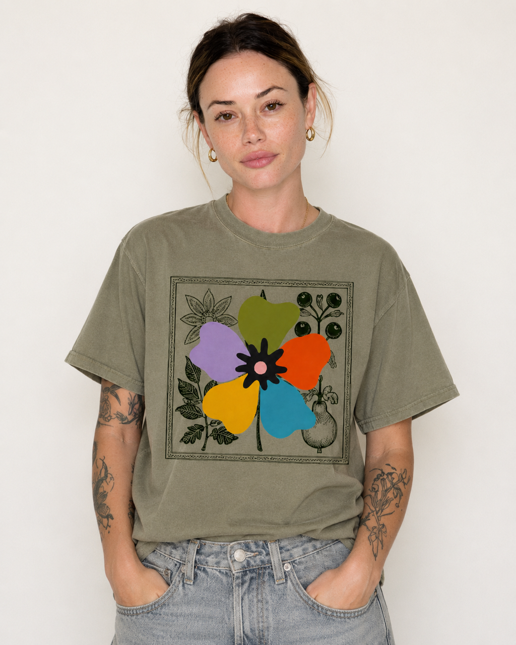 A woman wearing an olive green graphic t-shirt with a colorful floral print, paired with light-wash blue jeans, standing against a plain off-white background.