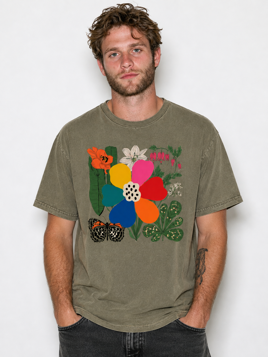 A man wearing a faded olive green t-shirt with a colorful graphic print of flowers, butterflies, and leaves, paired with dark gray jeans, against a plain light gray background.