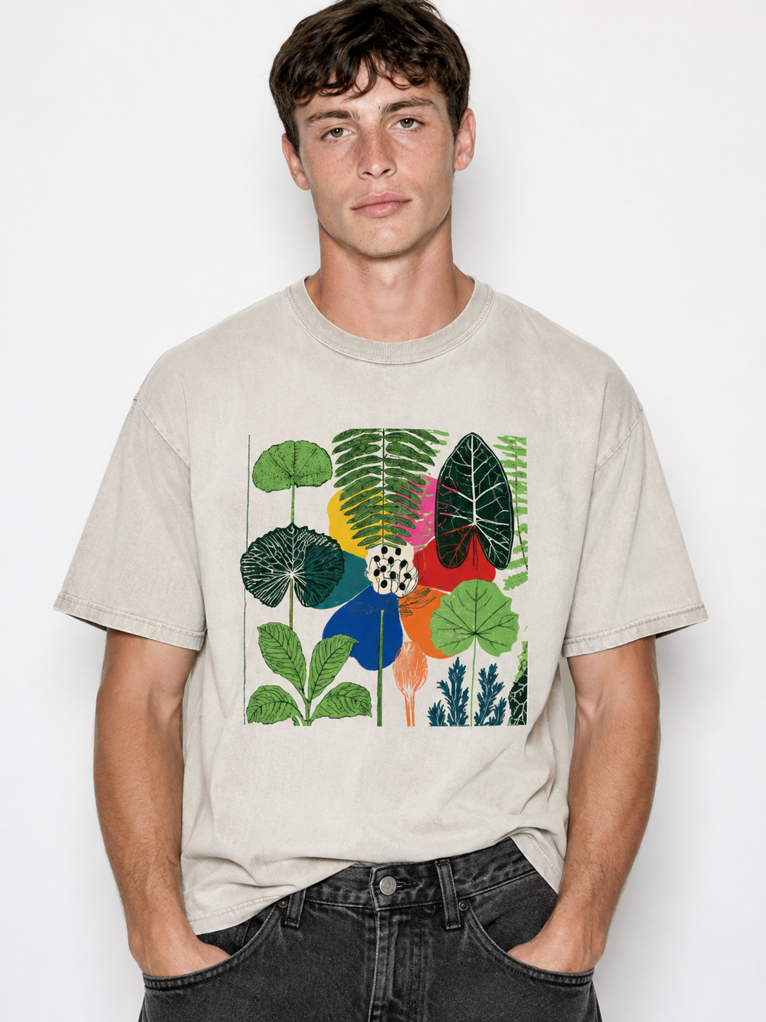 A man wearing a light beige crewneck t-shirt with a colorful tropical leaf graphic print on the front, paired with dark gray jeans, against a plain white background.