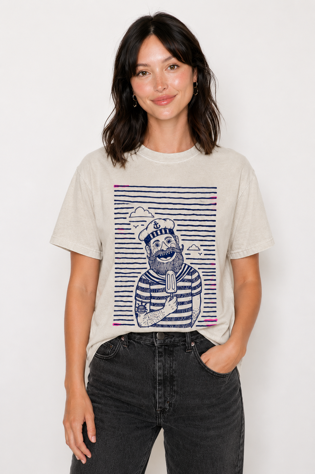 A woman modeling a light beige graphic t-shirt with a navy blue and pink striped cartoon sailor design, paired with black jeans, against a plain white background.