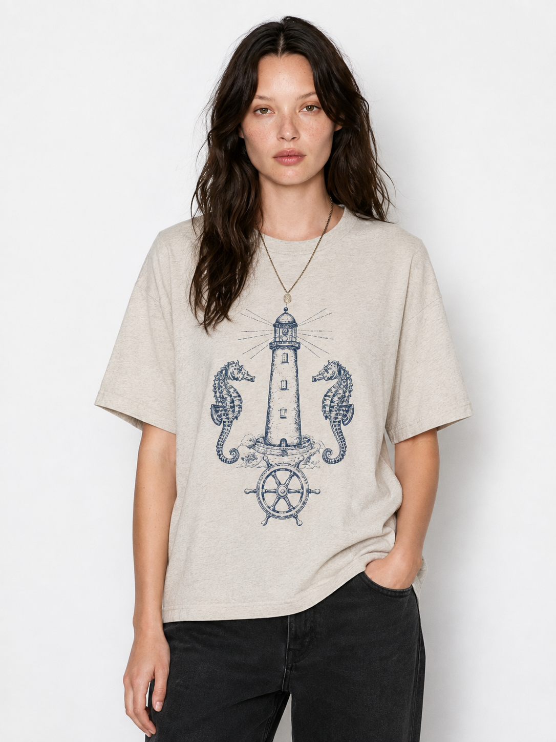 A woman modeling a light beige heather t-shirt with a navy blue graphic of a lighthouse, seahorses, and a ship's wheel, paired with black jeans against a white studio background.