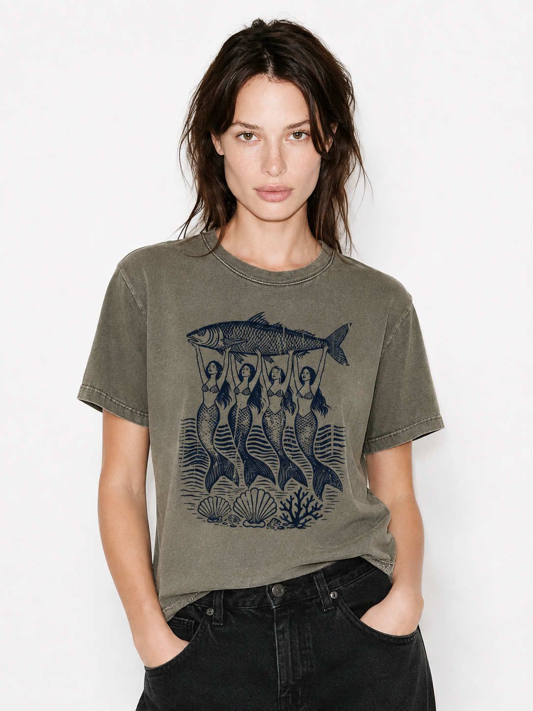 A woman wearing a washed olive green graphic t-shirt with a dark blue mermaid and fish design, paired with black jeans, against a plain white background.