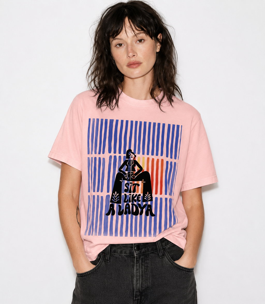 A woman wearing a light pink graphic t-shirt with a blue, orange, and black print that reads 'SIT LIKE A LADY' against a striped background, paired with dark gray jeans, set against a plain white studio background.