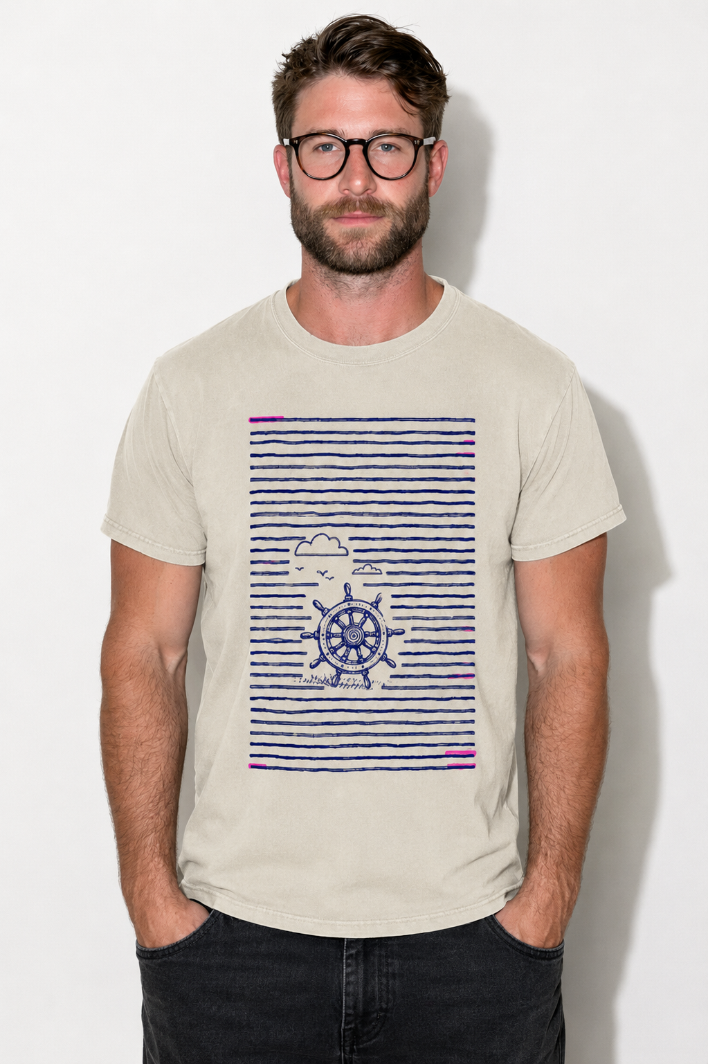 A man wearing a light beige crewneck t-shirt with a navy blue and purple nautical graphic on the front, paired with dark jeans, standing against a plain white wall.
