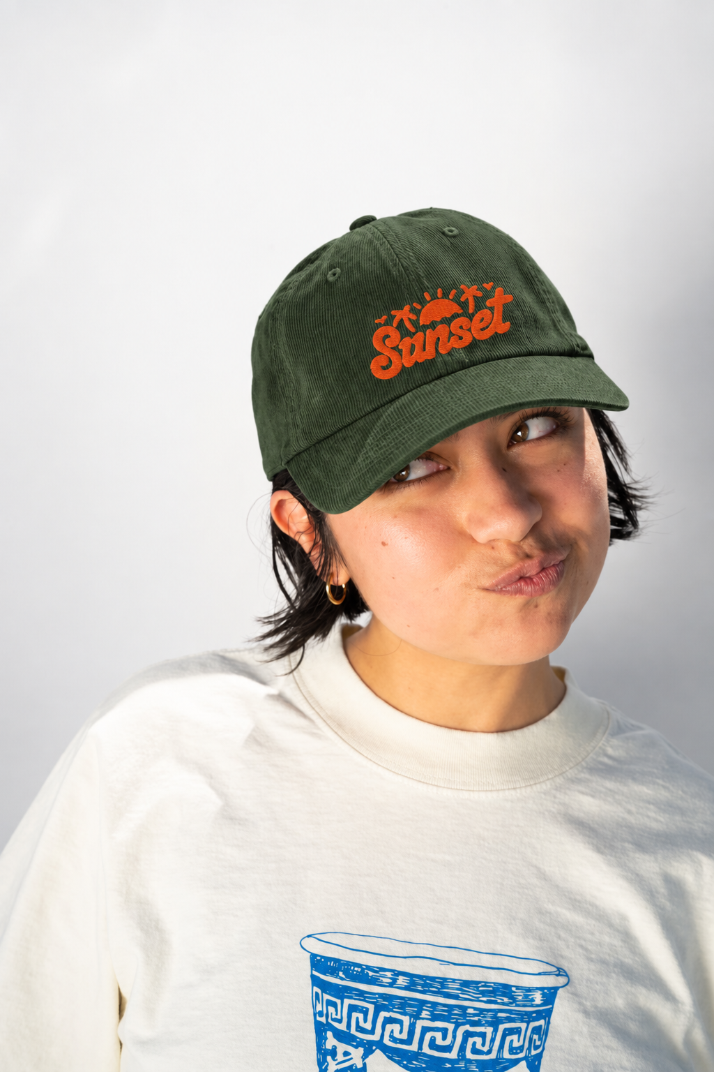 A person wearing a dark green baseball cap with orange embroidered 'Sunset' text and a white crewneck sweatshirt, photographed against a light gray studio background.