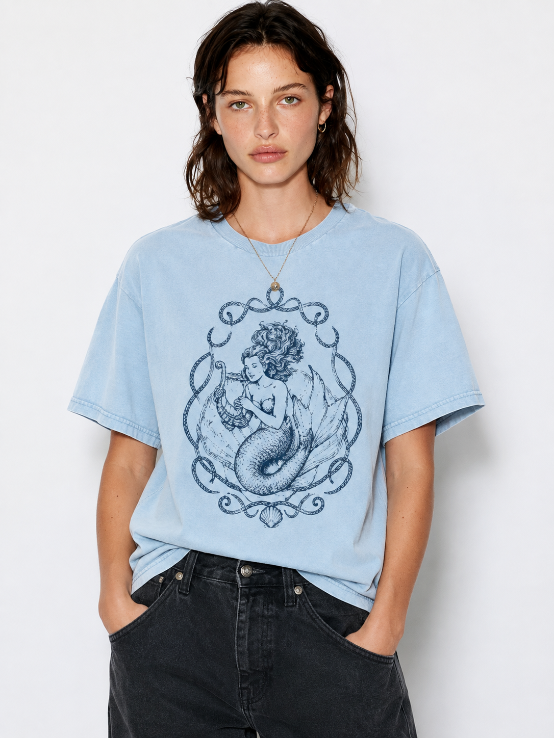 A woman wearing a light blue oversized graphic t-shirt with a dark blue mermaid illustration, paired with dark gray jeans, against a plain white studio background.