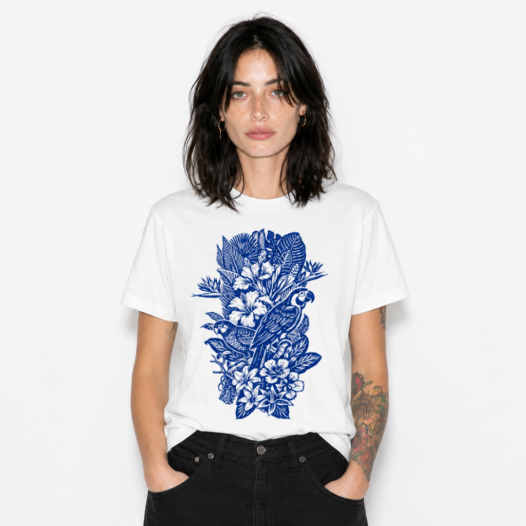 A woman modeling a white short-sleeve crewneck t-shirt with a blue tropical graphic print of a parrot, bird, and flowers on the front, paired with black jeans against a plain white background.