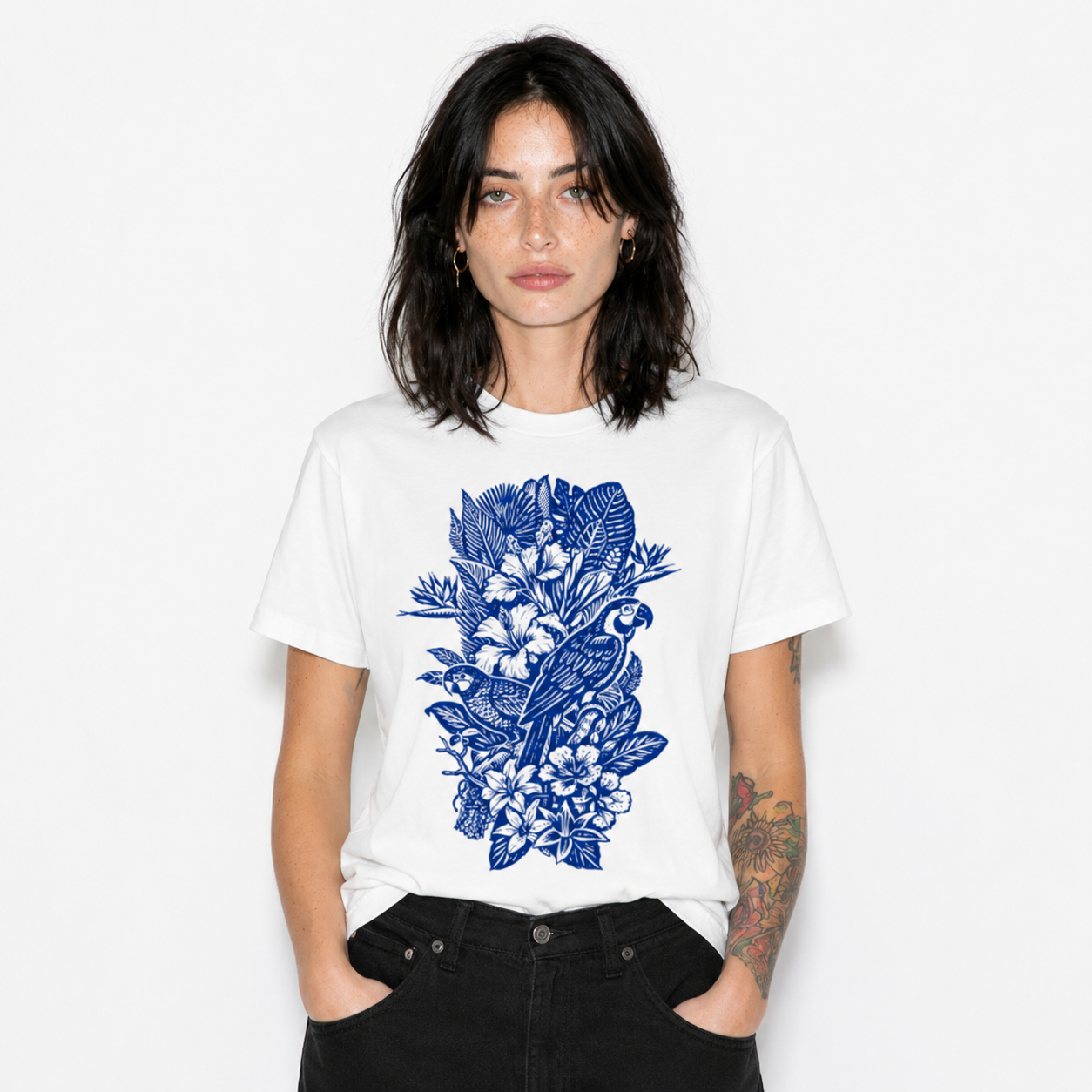 A woman modeling a white short-sleeve crewneck t-shirt with a blue tropical graphic print of a parrot, bird, and flowers on the front, paired with black jeans against a plain white background.