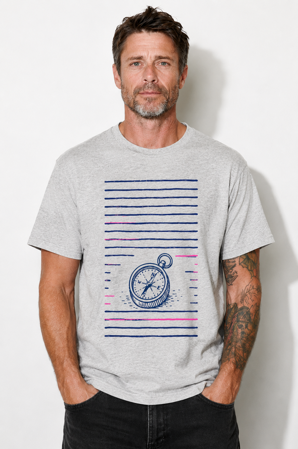 A man wearing a light gray heather crewneck t-shirt with a graphic print of a compass and horizontal blue and pink stripes, paired with black jeans, standing against a white wall.