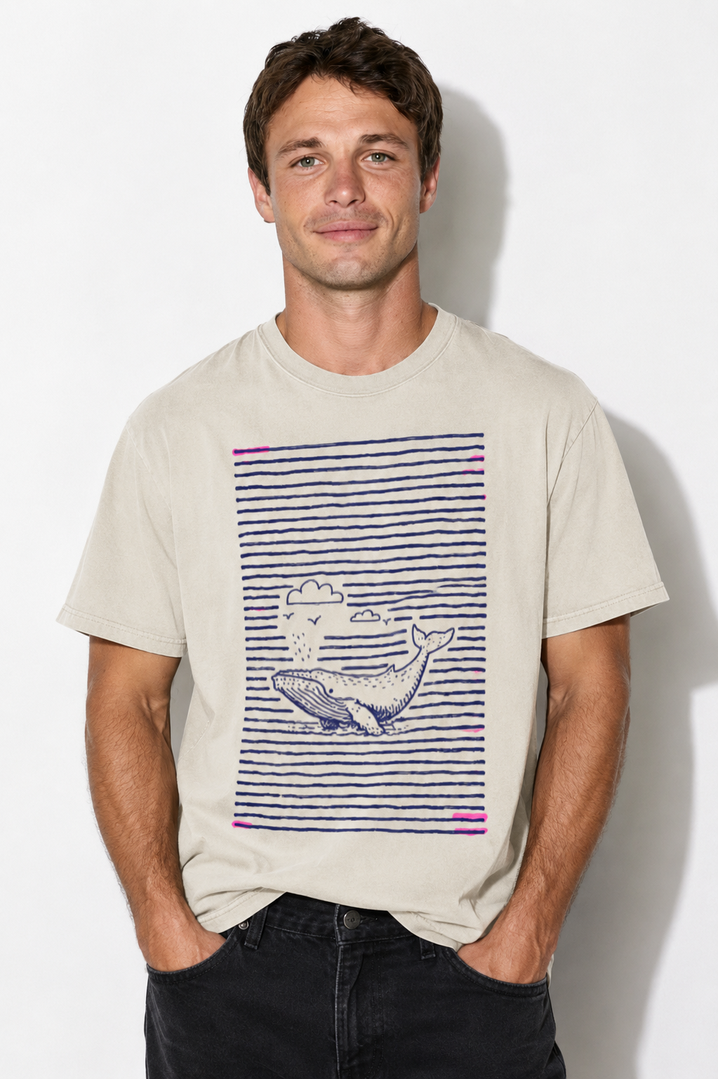 A man wearing a light beige t-shirt with a graphic print of a whale on a navy blue striped background, paired with black jeans, standing against a white wall.