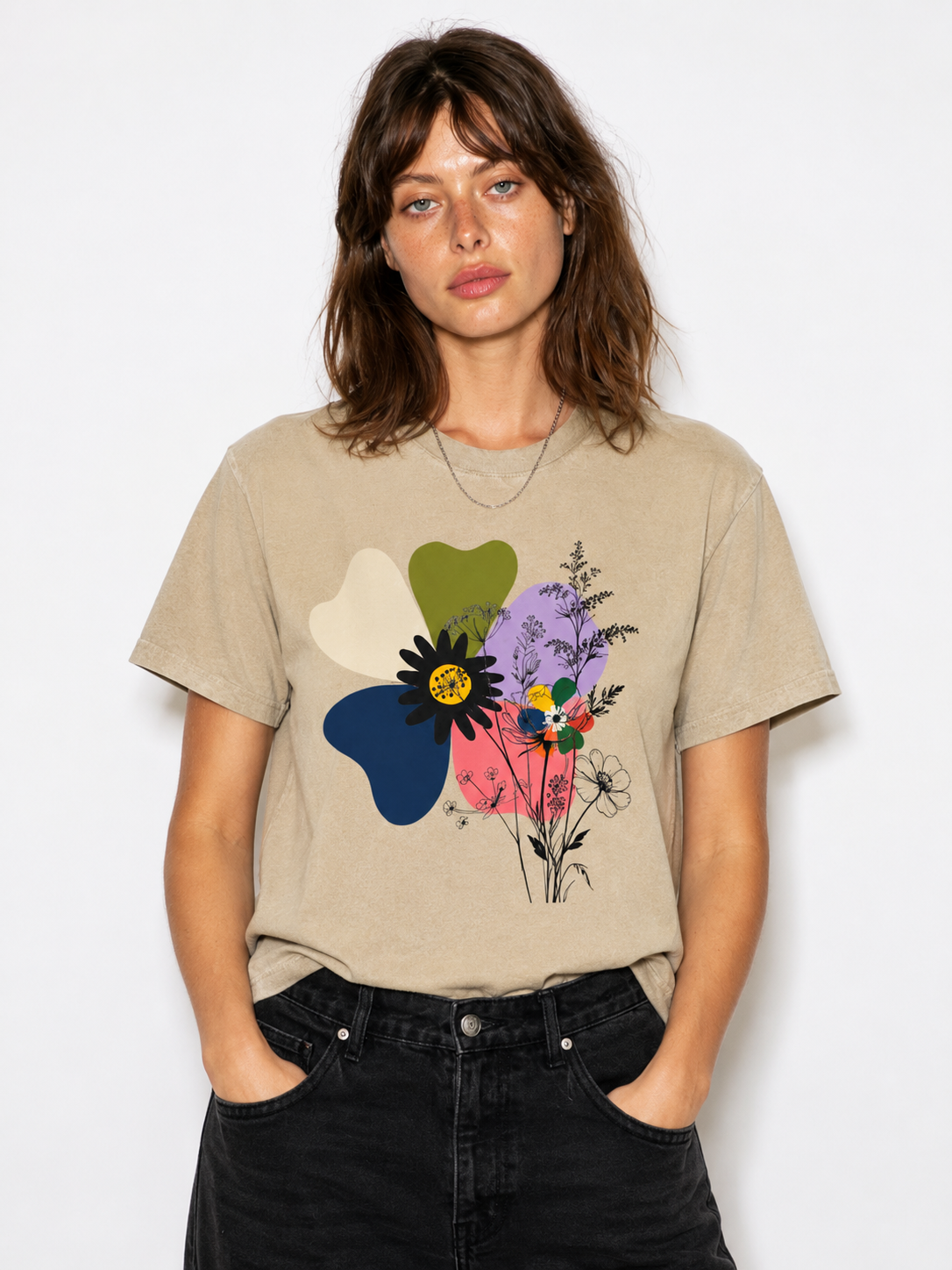 A woman wearing a beige graphic t-shirt with a colorful abstract flower design, paired with black jeans, against a plain white background.
