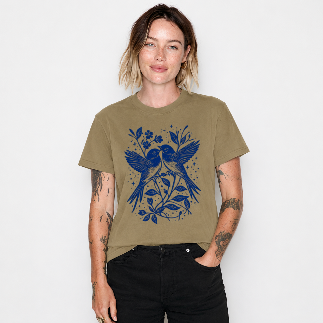 A woman wearing an olive green t-shirt with a blue bird and floral graphic print, paired with black jeans, standing against a plain white background.