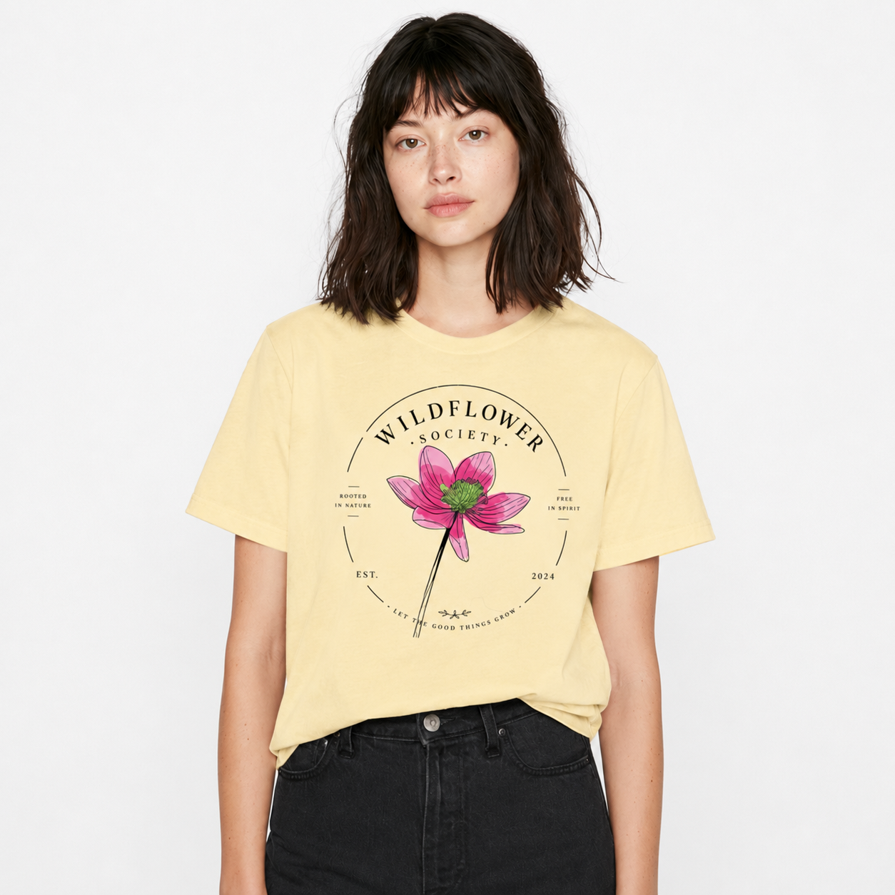 A woman wearing a pale yellow graphic t-shirt with a pink flower and 'Wildflower Society' print, paired with black jeans, against a plain white background.