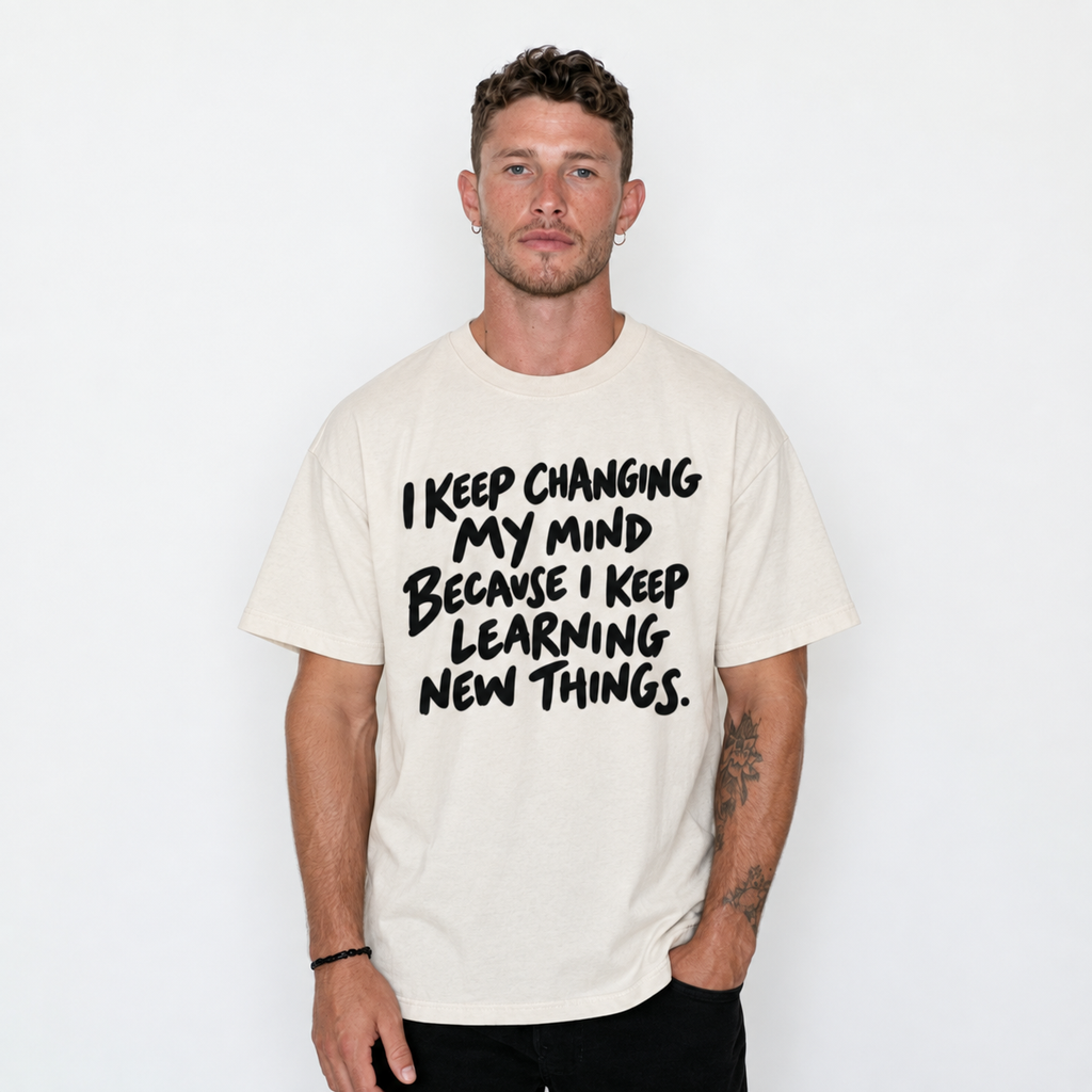 A man wearing a cream-colored t-shirt with black hand-drawn text that reads 'I KEEP CHANGING MY MIND BECAUSE I KEEP LEARNING NEW THINGS.' against a plain white background.